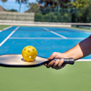 Close-up of a male hand holding a pickleball paddle and a yellow ball with holes