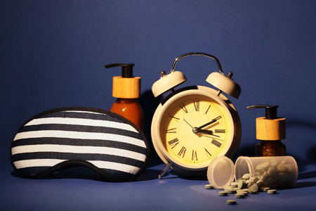 Sleeping mask, alarm clock, cosmetic bottles and pills on color background