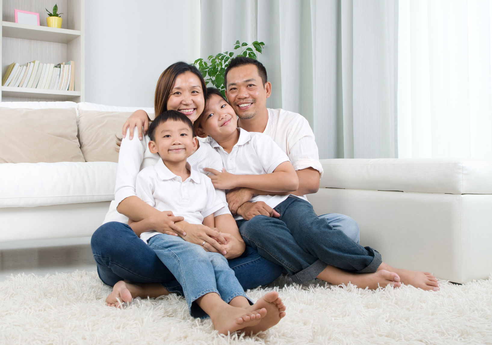 Asian family of four smiling at camera, sitting on living room carpet. Man, woman, and two boys