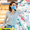 Woman in face mask choosing dairy products standing near shopping cart with yoghurt and milk