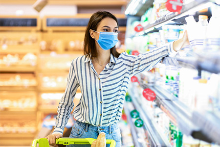 Woman in face mask choosing dairy products standing near shopping cart with yoghurt and milk