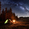 Night camping. Illuminated tent and campfire near forest under beautiful night sky full of stars and