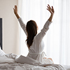 Young adult woman wear nightgown raising arms waking up happy concept in early good morning sitting 