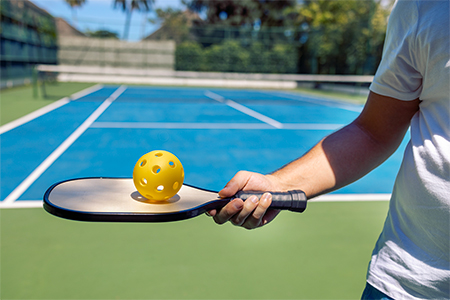 Close-up of a male hand holding a pickleball paddle and a yellow ball with holes