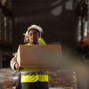 A female warehouse worker wearing protective workwear is carrying a box in a large warehouse