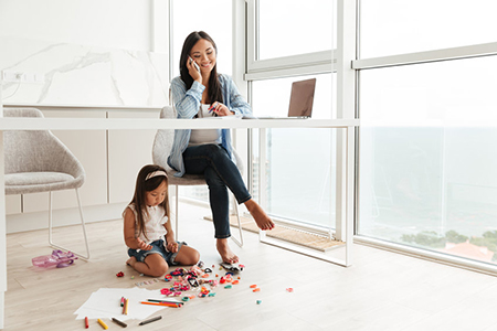 Young asian woman talking on a mobile phone and working on laptop while her little daughter playing 