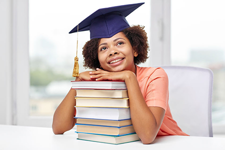  happy smiling african american student girl in bachelor cap with books sitting at table and dreamin