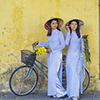 two women wearing long traditional Vietnamese dresses. holding bicycle in front of yellow wall