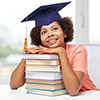  happy smiling african american student girl in bachelor cap with books sitting at table and dreamin
